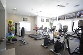 This fitness center is a thoughtfully designed, single-floor facility offering a comprehensive workout environment for users of all fitness levels. The space is organized into distinct zones, each catering to different exercise needs.

The main cardio area features a row of Matrix treadmills and elliptical trainers, positioned to face large windows that flood the room with natural light and views of the surrounding exterior. Ceiling fans overhead ensure comfortable air circulation during intense workouts. Multiple flat-screen TVs are mounted along the walls, providing entertainment throughout the exercise session.

A dedicated strength training zone houses a cable machine system, a free-weight dumbbell rack, and a weight bench, offering a well-rounded selection of resistance equipment. Potted greenery is placed throughout the space, adding a refreshing, natural element to the environment.

A separate cycling studio, enclosed by floor-to-ceiling glass walls, contains Matrix stationary bikes and an elliptical, maintaining an open and airy feel while providing a distinct workout zone. A water station is conveniently located nearby.

An additional room, visible through the glass partition, features a colorful kids' activity area with bright furnishings, making the facility family-friendly. Neutral-toned carpet, recessed lighting, and a clean, modern aesthetic tie the entire space together cohesively.