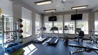 This fitness center is designed to provide a functional and comfortable workout environment. The space features dark rubber flooring, which offers durability and traction during exercise. Multiple windows line the walls, allowing abundant natural light to fill the room while offering views of the surrounding outdoor area, including what appears to be a pool or courtyard. Ceiling-mounted flat-panel lighting fixtures, along with a ceiling fan, ensure the space remains well-lit and properly ventilated. Several mounted TVs are positioned throughout the room for entertainment during workouts. The cardio equipment section includes multiple treadmills and at least one elliptical or stair climber machine. A weight rack along one wall holds an assortment of medicine balls and dumbbells in varying sizes, catering to both strength and conditioning workouts. The neutral gray walls contribute to a clean, modern aesthetic. The overall layout is thoughtfully organized, keeping cardio and strength equipment easily accessible while maintaining an open, uncluttered feel throughout the room.