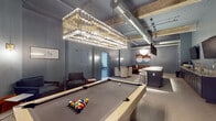 Located at 1801 Main St in Columbia, South Carolina, this single-floor amenity space blends industrial architectural character with refined, contemporary finishes throughout. Exposed concrete beams, visible ductwork, and soaring ceilings define the raw structural aesthetic, while carefully curated furnishings and lighting elevate the overall atmosphere into something polished and inviting.

The central gathering area is anchored by a full-size billiards table positioned beneath a dramatic crystal chandelier — a striking contrast of industrial and elegant elements. Surrounding the table, flexible lounge seating provides comfortable areas for socializing, complemented by curated artwork on the blue-gray walls that lend a cohesive, sophisticated palette to the space.

A built-in wet bar runs along one wall, featuring dark cabinetry with brass hardware, a richly veined marble countertop, an integrated sink with a brass faucet, and wall-mounted flat-screen televisions — ideal for entertaining. Adjacent lounge seating with upholstered chairs and a tufted sofa creates a more intimate gathering zone.

A dedicated fitness center, clearly marked and accessible from the main lounge area, houses cardio equipment including stationary bikes and an elliptical, a dumbbell rack with a weight bench, floor-to-ceiling mirrors, rubber flooring, and a cable resistance machine, providing a comprehensive workout environment within the same footprint.