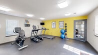Located at 20202 E Admiral Pl in Catoosa, Oklahoma, this fitness center offers a well-equipped workout facility across two connected rooms on a single floor. The main exercise area features durable rubber flooring and is highlighted by a bold yellow accent wall that adds energy to the space. Cardio equipment includes TRUE-brand treadmills, an elliptical trainer, and a stationary bike, while a wall-mounted flat-screen TV provides entertainment during workouts. A full rack of dumbbells lines one wall alongside a large mirror that visually expands the space and supports proper form during strength training. Additional functional fitness equipment includes a medicine ball rack and stability balls of varying sizes, complemented by workout instruction posters mounted on the walls. The secondary area features a glass-panel entry door that allows natural light to filter in, an adjustable workout bench, and additional wall-mounted signage. Flush ceiling light fixtures provide consistent overhead illumination throughout. The space maintains a clean, functional aesthetic with neutral white walls, gray trim, and a practical layout that accommodates a variety of exercise routines, making it a convenient and comprehensive fitness resource.