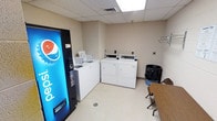 This single-floor shared laundry facility is designed for practical, everyday use. The room is finished with neutral beige walls and a drop ceiling with recessed fluorescent lighting, creating a bright and functional environment. The main feature of the space is a set of commercial-grade top-loading washing machines and front-loading dryers, arranged along the back wall and ready for resident use. A coin-operated or card-operated payment system is mounted above each machine, providing a straightforward self-service experience. A Pepsi vending machine is conveniently positioned near the entrance, offering refreshments while waiting for laundry to complete. A small table and chairs are also present, providing a comfortable spot to sit during the laundry cycle. The floor is finished with easy-to-clean vinyl tile, and a floor drain is centrally located for maintenance and safety purposes. A waste bin is accessible near the machines for added convenience. The overall layout is efficient and well-organized, with ample space to move around and manage laundry tasks with ease. This space is well-suited for a residential building, hotel, or similar multi-occupancy facility.