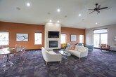 This well-appointed community clubroom offers a thoughtfully designed single-floor layout that seamlessly blends comfort and functionality. The expansive main lounge is anchored by a striking floor-to-ceiling tile-clad fireplace with a wall-mounted television, set against a warm terracotta accent wall. Vaulted ceilings with recessed lighting and a ceiling fan enhance the sense of openness, while large windows flood the space with abundant natural light and views of the surrounding landscape.

The lounge features multiple distinct seating arrangements, including contemporary white sofas paired with glass coffee tables and a separate nook furnished with teal accent chairs and a sculptural side table — ideal for small gatherings or quiet conversation. Patterned carpet tiles in a cool grey palette run throughout the social areas, adding texture and visual depth.

A dedicated transition zone with wooden arm chairs and direct glass door access leads out to a spacious outdoor deck, offering open-air views of the surroundings. The fully equipped kitchen impresses with rich dark espresso cabinetry, granite countertops, a mosaic tile backsplash, and a large central island with bar seating. Large-format stone-look tile flooring grounds the kitchen with a clean, modern aesthetic. Black appliances, including a refrigerator, range, and microwave, complete this functional and well-finished space.