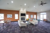 This well-appointed community clubroom offers a thoughtfully designed single-floor layout that seamlessly blends comfort and functionality. The expansive main lounge is anchored by a striking floor-to-ceiling tile-clad fireplace with a wall-mounted television, set against a warm terracotta accent wall. Vaulted ceilings with recessed lighting and a ceiling fan enhance the sense of openness, while large windows flood the space with abundant natural light and views of the surrounding landscape.

The lounge features multiple distinct seating arrangements, including contemporary white sofas paired with glass coffee tables and a separate nook furnished with teal accent chairs and a sculptural side table — ideal for small gatherings or quiet conversation. Patterned carpet tiles in a cool grey palette run throughout the social areas, adding texture and visual depth.

A dedicated transition zone with wooden arm chairs and direct glass door access leads out to a spacious outdoor deck, offering open-air views of the surroundings. The fully equipped kitchen impresses with rich dark espresso cabinetry, granite countertops, a mosaic tile backsplash, and a large central island with bar seating. Large-format stone-look tile flooring grounds the kitchen with a clean, modern aesthetic. Black appliances, including a refrigerator, range, and microwave, complete this functional and well-finished space.