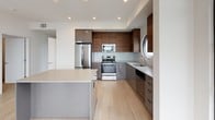 This single-floor residence offers a well-designed layout with two bedrooms and two full bathrooms, combining modern aesthetics with functional living spaces. The kitchen serves as a standout feature, showcasing a blend of warm walnut-toned upper cabinetry and sleek gray lower cabinets, complemented by a light quartz countertop and a distinctive circular porthole window above the sink. A full suite of stainless steel appliances — including a French door refrigerator, range, microwave, and dishwasher — is integrated seamlessly into the design. A central island provides additional workspace and casual seating. Light wood-toned flooring runs throughout the main living areas, enhancing the open and airy feel of the space. The bedrooms are carpeted for comfort, each benefiting from large windows with exterior views that bring in natural light. Both bathrooms are finished with tile flooring, wood-toned vanity cabinetry, and full tub-and-shower combinations accented by horizontal tile surrounds. One bedroom includes an adjacent walk-in closet, adding practical storage. Recessed lighting throughout reinforces the clean, contemporary ambiance of the residence.