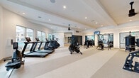 Located at 2045 Paseo Belluno in Chula Vista, CA, this modern fitness center offers a thoughtfully designed, single-floor layout with ample space for a comprehensive workout experience. The main floor is defined by high, coffered ceilings with recessed lighting and ceiling fans that enhance airflow and comfort throughout. A durable, speckled rubberized floor in a subtle striped pattern covers the primary workout area, providing both functionality and a clean aesthetic.

The cardio zone lines one side of the main floor, featuring a row of TRUE-brand treadmills, air bikes, and stair climbers positioned near large windows that invite natural light and outdoor views. Opposite, a strength training section showcases a robust selection of TRUE selectorized machines, free weight benches, a cable system, and a well-stocked dumbbell rack, all reflected in full-length framed mirrors mounted along the walls. Wall-mounted flat-screen TVs keep members engaged during workouts.

A dedicated TRX functional training studio is separated from the main floor and features warm hardwood flooring, a full-wall mirror with a ballet barre, a TRX suspension training rig, and curated fitness wall art — creating an intimate space ideal for group or individual training. A serene watercolor mountain mural accents a transitional wall near the strength area, adding an artistic touch to the overall ambiance. The space is modern, clean, and purposefully equipped for diverse fitness needs.