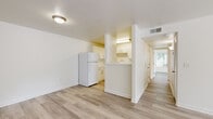 This single-floor residence offers a thoughtfully designed layout comprising two bedrooms, two bathrooms, a living room, kitchen, and hallway. Light-toned wood-style flooring runs consistently throughout, lending a clean, cohesive look to the entire space.

The open living area is bright and airy, with neutral white walls and natural light filtering in through windows that offer views of surrounding greenery. A paneled entry door anchors one end of the living space, while a clear sightline connects to the kitchen area.

The kitchen features an L-shaped layout with light-speckled stone countertops, white cabinetry equipped with sleek bar-pull hardware, and a stainless steel sink. A full-size refrigerator is positioned adjacent to the kitchen, visible from the living area.

The primary bedroom is generously proportioned, with a large window that fills the room with natural light and garden views. Both bedrooms include closets with built-in shelving rods for organized storage.

The two bathrooms share a consistent design language, each featuring a dark navy vanity with a white basin sink, a framed mirror with overhead lighting, wood-style flooring, and a tiled shower area. The overall ambiance of the residence is clean, modern, and move-in ready, with a neutral palette that provides a versatile backdrop for any interior style.