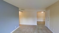 This single-story space presents a clean, functional layout with a straightforward design aesthetic. The main living area features wall-to-wall carpeting in a neutral beige tone, complemented by white baseboards and a combination of white and soft blue-gray accent walls that add a subtle visual contrast to the otherwise understated palette. A textured ceiling adds a layer of detail overhead, while HVAC vents are integrated throughout the ceiling for climate control.

An arched doorway serves as the architectural focal point of the space, connecting the carpeted main living area to an adjacent room finished with hard surface flooring. This secondary area is equipped with a ceiling fan, providing both lighting and air circulation. The arched transition gives the layout a sense of continuity while visually distinguishing the two areas.

The walls are clean and move-in ready, offering a blank canvas for customization. Natural light appears to supplement the interior lighting, contributing to the bright, open feel of the space. Overall, the layout is practical and well-organized, suitable for a variety of uses across its connected living zones.
