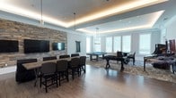 This single-floor entertainment space is thoughtfully designed to serve as a premier gathering and recreation area. The room immediately draws the eye with a striking stacked-stone accent wall anchoring a large flat-screen television, creating a dedicated media and lounge zone complete with a long bar-height communal table and cushioned high-back chairs — ideal for viewing events or casual socializing.

The space transitions seamlessly into a game area, where a full-size billiards table takes center stage beneath an elegant pendant light fixture, accompanied by a foosball table set atop a decorative area rug. Natural light pours in through large windows along the far wall, brightening the open layout and complementing the warm-toned hardwood flooring underfoot.

Architectural interest is added by a coffered tray ceiling with integrated ambient LED strip lighting, lending the room a polished, upscale character. Recessed ceiling lights provide additional illumination throughout. The neutral color palette of soft grey walls and light wood floors creates a clean, modern aesthetic that balances comfort with sophistication. Overall, the space offers a versatile and well-equipped environment for entertainment, recreation, and community engagement.