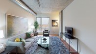 This single-floor unit showcases a distinctive industrial-loft aesthetic defined by exposed concrete ceilings and visible ductwork running throughout the space. The living area is anchored by large windows that frame urban views of the surrounding cityscape, allowing generous natural light to fill the room. Hardwood flooring and concrete structural columns complement the raw, urban character of the space.

The kitchen is well-appointed with rich dark-wood cabinetry, granite countertops, under-cabinet lighting, and a full suite of stainless steel appliances, including a range, microwave, dishwasher, and refrigerator. A pass-through opening connects the kitchen to adjacent living areas, reinforcing the open feel.

Two bedrooms feature carpeted floors and exposed concrete ceilings consistent with the loft aesthetic, each with windows providing natural light. One bedroom includes direct access to an en-suite bathroom.

Both bathrooms are finished with dark-tile flooring, granite-topped vanities with dark wood cabinetry, and large mirrors. One bathroom features a tub-shower combination, while the other offers a walk-in shower, providing functional variety throughout the unit.