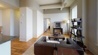 This single-floor residence offers a compelling blend of historic architectural character and contemporary comfort. The open living area is anchored by rich hardwood flooring, exposed ceiling beams, and a classic fireplace, creating a warm and inviting atmosphere. Tall windows in the living room frame city views and flood the space with abundant natural light, complementing the neutral palette of warm beige and greige tones found throughout.

The kitchen is well-appointed with warm wood cabinetry, a tile backsplash, generous counter space, and recessed lighting overhead. Hardwood floors continue seamlessly into the adjacent dining area, where tall windows provide additional natural light and urban views, making it an ideal spot for everyday dining.

The bedroom features the same warm hardwood floors and large windows that allow ample daylight in, supported by crown molding details that add architectural refinement. A second, more intimate bedroom provides a cozy retreat with soft lighting.

The primary bathroom is equipped with a wide vanity, dual sinks, wood cabinetry, and tile flooring, offering practical storage and a clean aesthetic. A secondary bathroom features a classic pedestal sink with dark tile flooring, adding a touch of timeless style. Together, the rooms create a cohesive, well-designed living environment with notable historic urban character.