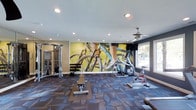 This single-floor fitness space is designed to support a well-rounded workout experience. The room features a combination of strength and cardio equipment, including a multi-function cable machine, a weight bench, a dumbbell rack, and stationary cycling bikes positioned near a window. A striking large-scale wall mural depicting a close-up of a cyclist in motion serves as a bold focal point, energizing the space with a dynamic, motivational aesthetic. The patterned blue-grey carpet adds texture underfoot while defining the workout area. Recessed lighting throughout the ceiling provides consistent, even illumination, complemented by a ceiling fan that aids in air circulation during exercise. A window on the far wall allows natural light to filter into the space and offers a glimpse of outdoor greenery, contributing to an open, airy feel. The walls are finished in contrasting tones, with a deep teal-grey accent wall framing the window area and lighter neutral tones elsewhere, creating visual depth. Overall, the space offers a focused and functional environment suited for both cardio and resistance training.