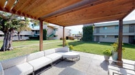 The pergolas at 2904 Clairemont in San Diego's Clairemont neighborhood serve as a well-designed communal outdoor retreat within a residential community. The centerpiece is a generously sized pergola structure, constructed with rich warm-toned wood beams and a solid tongue-and-groove cedar ceiling, providing ample shade while maintaining an open, airy feel. Beneath the pergola, a poured concrete patio hosts contemporary modular outdoor seating, including a large curved sectional sofa and coordinating lounge chairs and coffee tables in neutral tones, creating a comfortable and stylish gathering space.

The pergola opens directly onto an expansive, well-maintained lawn that forms the heart of the community courtyard. Landscaped planting beds featuring ornamental grasses, succulents, and decorative rock borders line the pathways, adding texture and visual interest throughout. Concrete walkways cut cleanly through the greenery, connecting various areas of the grounds. Mature trees provide additional shade and greenery throughout the outdoor space.

Views from the pergola look out across the manicured courtyard toward the surrounding two-story residential buildings, with a glimpse of a pool area visible in the distance. The overall ambiance is relaxed and inviting, offering residents an attractive outdoor environment suited to San Diego's sunny climate.