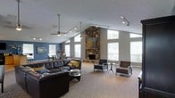 This single-floor space is defined by its striking architectural character and open, airy layout. The vaulted ceilings rise dramatically overhead, creating a generous sense of vertical space that is further enhanced by an abundance of natural light streaming through large windows along the walls. At the heart of the room stands a floor-to-ceiling stone fireplace, serving as a bold focal point that anchors the space with rustic elegance. Track lighting and a ceiling fan are thoughtfully installed to complement both function and ambiance. The main living area features a large, dark leather sectional sofa with accent pillows, paired with modern armchairs and side tables arranged to encourage conversation and relaxation. A glass-top coffee table adds a contemporary touch. The neutral carpet flooring and taupe-toned walls are accented by a rich blue accent wall, lending a sense of warmth and color depth. Toward the rear, a separate dining area is visible, with seating for multiple guests, making this space well-suited for both casual gatherings and more structured events. Decorative artwork adorns the walls, contributing to an inviting and polished atmosphere throughout.