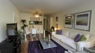 This single-floor residence offers a thoughtfully designed open-concept layout that seamlessly connects the living, dining, and kitchen areas into one cohesive and functional space. The living area is anchored by a plush white sofa, a sleek mirrored coffee table, and a deep-toned area rug that adds warmth and contrast to the rich hardwood flooring that runs throughout the space. Decorative accents, including framed wall art and lush indoor plants, contribute to a refined yet inviting ambiance. A ceiling fan with integrated lighting serves as a practical centerpiece overhead, providing both airflow and ambient illumination. The dining area accommodates a contemporary table with modern white chairs, positioned near a stylish open shelving unit that adds a decorative and functional touch. Adjacent to the dining space, a pass-through area connects to the kitchen, enhancing the open feel of the floor plan. Warm accent lighting from a floor lamp adds a cozy glow to the space in the evenings. The overall aesthetic blends modern furnishings with warm tones and natural textures, creating a comfortable and visually appealing environment well-suited for both everyday living and entertaining.