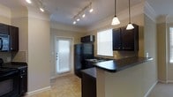 This single-floor space offers a thoughtfully designed interior that balances functionality with style. The kitchen serves as a central highlight, featuring rich dark wood cabinetry that contrasts beautifully with dark granite-style countertops. A peninsula breakfast bar extends the counter space, providing an ideal spot for casual dining or entertaining. Black appliances, including a full-size refrigerator and built-in microwave, complement the kitchen's cohesive aesthetic. Pendant lighting hangs gracefully above the bar, while a track lighting system illuminates the kitchen and adjacent areas with adjustable, directed light. Crown molding frames the ceilings throughout, adding a refined architectural detail to the space. Natural light filters generously through multiple windows fitted with white horizontal blinds, creating a bright and welcoming ambiance. Light-toned tile flooring runs through the main areas, providing a clean and durable foundation. A door with a full-length window panel offers direct access to the exterior, further enhancing the sense of openness and connectivity to the outdoors. Overall, the space presents a well-maintained, move-in-ready environment with a polished and practical design.
