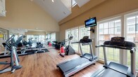 Located at 5353 Wilcox Rd in Dublin, Ohio, this fitness center offers a spacious and well-appointed workout environment designed to support a full range of exercise routines. The facility is characterized by soaring vaulted ceilings that create an open, airy atmosphere, complemented by warm beige walls and rich hardwood-style flooring throughout.

Natural light pours in through large, multi-pane windows lining the walls, providing views of the surrounding greenery and keeping the space bright and inviting. Full-length mirrors are strategically placed to enhance the sense of space and assist exercisers with form and technique.

The cardio section is well-stocked with Life Fitness treadmills, elliptical trainers, and a stationary recumbent bike, all positioned to take advantage of the natural light and outdoor views. A wall-mounted flat-screen television provides entertainment during workouts. The strength training area features a multi-station cable machine, a weight bench, and a comprehensive set of dumbbells on tiered racks. Stability balls and additional functional training accessories round out the free weights area.

An upper-level mezzanine overlooks the main floor, adding architectural interest to the space. Glass partitions near the entrance offer a glimpse into an adjoining lounge or common area. The overall ambiance is clean, modern, and energizing, making this fitness center a functional and comfortable space for daily exercise.