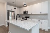 This single-story, one-bedroom, one-bathroom unit in Advance, NC is thoughtfully designed with a clean, contemporary aesthetic throughout. The open-concept layout connects the living room and kitchen, creating a cohesive and functional living space. Light wood-tone flooring runs through the main areas, reflecting natural light and contributing to a bright, airy atmosphere. Ceiling fans are present in both the living room and bedroom, adding comfort and practicality.

The kitchen is a standout feature, showcasing white shaker-style cabinetry with matte black hardware, granite countertops, a tile backsplash, and a central island with pendant lighting and an undermount sink. Stainless steel appliances, including a refrigerator, range, and over-the-range microwave, complete the space. The living area includes door access to the exterior, allowing natural light to pour in through slatted blinds.

The bedroom is carpeted for comfort and features a ceiling fan, windows with blinds, and access to a walk-in closet equipped with multi-tiered wire shelving. The bathroom mirrors the home's design language with a white vanity, granite countertop, and wood-tone flooring. Built-in open shelving adds convenient storage nearby.