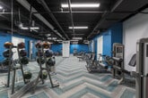 This fitness center in Decatur, Georgia, offers a spacious, single-floor layout designed to accommodate a comprehensive range of workout activities. The interior showcases a striking industrial aesthetic, with an exposed black ceiling that houses rows of bright linear LED lighting, ensuring the space is consistently and evenly illuminated throughout.

Bold teal accent walls punctuate the otherwise white walls, adding an energetic visual contrast that reinforces the facility's active atmosphere. Underfoot, a distinctive chevron-patterned carpet in neutral tones with teal accents provides both visual interest and a practical, durable surface across the open floor plan.

The facility is thoroughly equipped with a wide selection of cardio machines, including ellipticals, stationary spin bikes, and rowing equipment, all arranged to maximize usable floor space. A dedicated strength-training area features cable and resistance machines, while a free-weight section offers a full rack of dumbbells alongside adjustable benches. Medicine balls and additional functional training tools round out the available equipment.

Full-length mirrors along one wall visually expand the space and serve a practical purpose for form monitoring during workouts. The overall layout promotes an efficient flow between exercise zones, making this fitness center a well-organized and functional training environment.