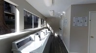 This shared laundry facility is a bright, functional space designed to accommodate multiple users efficiently. A long row of top-loading washing machines lines the wall beneath a series of large windows, allowing generous natural light to fill the room and create an open, airy atmosphere. The windows also offer views to the exterior, adding a sense of connection to the outdoors. Dryers are positioned along the opposite wall, providing a streamlined workflow for laundry tasks. The room features dark hardwood-style flooring that contrasts cleanly with the light grey walls and white appliances, giving the space a modern, tidy appearance. A textured ceiling with overhead lighting ensures the room remains well-lit at all times. Instructional signage is posted on the walls, offering clear guidance on machine usage and laundry procedures. Storage and utility panels are neatly integrated into the space. The overall layout is practical and efficient, making excellent use of the available square footage to serve the needs of building residents in a clean and orderly environment.
