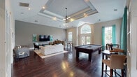 Located at 1000 Glen Forest Drive in Greenville, South Carolina, this single-floor clubhouse offers a well-appointed communal gathering space defined by refined architectural design and versatile amenities. The interior is anchored by rich, dark hardwood flooring that runs throughout, complementing the neutral gray and warm beige wall tones that lend a sophisticated, cohesive aesthetic.

The main lounge area features soaring vaulted and coffered ceilings, creating a generous sense of volume and openness. Large grid-pane windows and glass-paneled French doors flood the space with natural light while framing views of the outdoor pool area, seamlessly connecting the interior to the exterior. Decorative arched doorways supported by classical columns divide zones gracefully, adding architectural character.

The game room accommodates a full-size billiard table alongside intimate bistro-style seating beneath tall windows dressed in vibrant teal drapery. A comfortable lounge zone features a plush sectional sofa, accent chairs, and a wall-mounted television, ideal for relaxed gatherings. A beverage station is thoughtfully integrated into the layout. The entry area welcomes visitors with a patterned rug, accent seating, and fresh floral arrangements beneath the soaring ceiling. Throughout, curated artwork and carefully selected furnishings contribute to an inviting, polished ambiance.