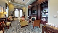 This single-floor space functions as a well-appointed communal gathering area designed to balance comfort with practicality. The main lounge is anchored by a rich, dark-wood built-in wall unit housing a fireplace, a mounted television, and flanking bookshelves stocked with reading materials. Comfortable sofas and upholstered armchairs are arranged around a glass-topped coffee table, creating an inviting seating area ideal for relaxation or casual conversation. Tall arched windows dressed with tropical-print valances allow generous natural light to fill the room, complementing the soft sage-green walls and crisp white crown molding. A tray ceiling adds architectural dimension and a sense of heightened volume to the space.

An adjoining area serves as a business or study nook, equipped with a work desk, computer station, and a cozy reading chair beside a tall window. A separate service counter features white cabinetry, a bulletin board, and a beverage station, providing functional amenities for guests or residents. Throughout the entire space, a bold navy and tan geometric-patterned carpet ties each zone together cohesively, while warm lamp lighting enhances the overall inviting and refined ambiance.
