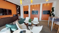 This single-floor lounge space makes a strong visual impression through its bold design choices and carefully curated furnishings. The most striking feature is the vivid orange accent walls, which are recessed into architectural niches and illuminated by recessed spotlights, creating a warm and energetic ambiance throughout the room. White trim and molding contrast sharply against the orange, lending the space a clean, polished look.

The seating area is centered around a circular glass-top table with a chrome base, surrounded by sleek, modern white swivel chairs with a sculptural silhouette. A small potted succulent serves as a minimalist centerpiece. Adjacent to this grouping, teal upholstered lounge chairs add a complementary pop of color and additional comfort.

Multiple flat-screen televisions are mounted across the orange-accented walls, making the space well-suited for presentations, entertainment, or collaborative use. A dark wood built-in shelving unit along one wall provides functional storage and display space, while a small credenza with office equipment suggests a multipurpose function. Pendant lights with cylindrical amber shades hang from the white ceiling, reinforcing the warm color palette. Wood-look flooring ties the entire space together with a grounded, contemporary feel.
