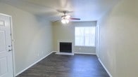 This single-floor residence offers a clean, functional layout designed for comfortable everyday living. The living area serves as the heart of the space, anchored by a classic fireplace with a white surround that adds a warm, inviting focal point to the room. Dark hardwood flooring extends throughout, providing a rich contrast against the light, neutral-toned walls that lend the space an open and airy feel. A multi-blade ceiling fan with an integrated light fixture is centrally positioned overhead, ensuring year-round comfort and ample illumination. Natural light flows in through a large window dressed with horizontal blinds, brightening the interior and connecting the living space to the outdoors. The clean white baseboards and smooth wall finishes contribute to the overall tidy and well-maintained character of the space. The thoughtful combination of practical features — from the fireplace to the hardwood floors — creates a cohesive and welcoming atmosphere throughout this single-story home.