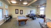 This single-floor recreation lounge is thoughtfully designed to serve as a social hub, combining game-room functionality with a comfortable, relaxed atmosphere. The centerpiece of the space is a full-size pool table with a blue felt surface and classic wooden frame, positioned prominently to allow ample room for play on all sides. The carpeted flooring adds warmth underfoot and helps define the space's inviting character.

A tray ceiling with recessed edges adds architectural interest and a sense of height to the room, while ceiling fans ensure comfortable air circulation throughout. Flush-mounted light fixtures provide even, bright illumination across the space.

Adjacent to the game area, a well-appointed lounge seating arrangement features a dark leather sofa, upholstered armchairs, a coffee table, and coordinating end tables with table lamps, creating an ideal spot for conversation or relaxation. Framed black-and-white artwork adorns the walls, adding a refined touch to the décor.

Overall, the space strikes a balance between casual recreation and comfortable lounging, making it well-suited for community gatherings and social interaction.