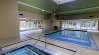 This indoor pool facility is housed within a single-story structure distinguished by its striking vaulted ceiling, which rises to a prominent peak and is fitted with a large central skylight. The skylight floods the pool area with natural light, creating an open, airy atmosphere despite the fully enclosed setting. The ceiling's angular framework is accented by a green finish along the upper panels, adding a subtle architectural detail that contrasts with the neutral, warm-toned walls below. Pendant light fixtures supplement the natural illumination, ensuring consistent brightness throughout the space. The rectangular pool itself is the focal point of the room, featuring clear blue water and stainless steel handrails for safe entry and exit. A wide tiled surround borders the pool on multiple sides, providing ample deck space for swimmers and guests. Along the back wall, large windows allow additional light to filter in, while a recessed alcove and bench seating offer a functional rest area. The overall ambiance is clean, functional, and inviting, making this facility well-suited for both recreational swimming and structured aquatic activities.