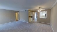 This single-floor residence offers a practical and comfortable layout across two bedrooms, two bathrooms, and a dedicated den. The main living area is generously proportioned, featuring neutral-toned carpeting, crown molding, and warm taupe walls that create a cohesive and inviting atmosphere. A ceiling fan with integrated lighting fixture serves as a functional centerpiece overhead.

A standout built-in cabinetry unit divides the living space, featuring glass-front upper cabinets and lower storage below, providing both display and organizational functionality. This architectural detail adds character while enhancing the room's utility.

Natural light filters in through windows visible toward the back of the space, brightening a hallway that connects to additional rooms. A pass-through or windowed area adjacent to what appears to be a kitchen area allows light to flow between spaces. The den provides flexible additional space suitable for a home office or guest area. Doorways lead to the bedrooms and bathrooms, completing a well-organized, single-level floor plan designed for everyday livability.