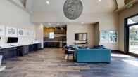 Located in Saint Petersburg, Florida, this single-floor community clubhouse at Brighton Bay offers a thoughtfully designed gathering space that blends modern aesthetics with functional amenities. The interior is anchored by rich wood-look tile flooring that flows seamlessly throughout, complementing the neutral warm-toned walls and crisp white ceilings.

Distinctive arched architectural details frame the open-plan layout, with structural columns adding a refined, Mediterranean-inspired character. Soaring ceilings amplify the sense of openness, while recessed lighting and a striking spherical pendant light fixture serve as focal design elements.

The social lounge area features plush teal sectional seating and a mounted flat-screen television, with floor-to-ceiling windows and glass doors that invite abundant natural light and provide direct views of the outdoor pool area. An adjacent seating nook with vibrant yellow accent chairs and a round coffee table offers an inviting spot to relax.

The kitchenette is outfitted with dark cabinetry, granite countertops, open shelving, a stainless steel sink, a wine refrigerator, and eye-catching yellow tile accents on the backsplash. A dedicated business center provides a built-in granite-topped workstation with computer access and printing capabilities. A dining area with a large table and modern chairs completes the versatile, community-focused layout.