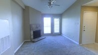 This single-floor residential space offers a comfortable and functional layout designed for everyday living. The main living area serves as the heart of the home, highlighted by a classic brick fireplace with a raised hearth that adds warmth and character to the room. Soft wall-to-wall carpeting runs throughout, contributing to a cozy and inviting atmosphere. A ceiling fan with an integrated light fixture is centrally mounted, providing both air circulation and ambient lighting. Natural light flows generously into the space through a large window and an adjoining glass-paneled exterior door, which together brighten the interior and create a seamless visual connection to the outdoors. The neutral-toned walls provide a versatile backdrop that complements a variety of décor styles. Clean white baseboards frame the room with a crisp finish, while the overall layout offers an open and unobstructed feel with ample room to furnish and personalize. The single-story design ensures easy accessibility throughout, making this a practical and welcoming space suited for a range of living arrangements.