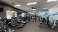 This single-floor fitness center is thoughtfully designed to support a comprehensive workout experience. The space is outfitted with a generous array of cardio equipment, including multiple treadmills, elliptical machines, and stationary bikes, all arranged to maximize floor space and ease of movement. A dedicated strength training zone features cable machines, a weight bench, and a fully stocked dumbbell rack, catering to a wide range of fitness routines.

One of the most visually distinctive features is a large, vibrant water splash mural that spans an entire wall, adding an energetic and dynamic aesthetic to the space. Full-length mirrors line one side of the room, visually expanding the area and providing practical utility for form-checking during workouts. Overhead, flush panel lighting and ceiling fans ensure the space remains well-lit and comfortable throughout use.

Windows along the exterior wall allow natural light to filter in and offer views of an adjacent outdoor pool area, creating a bright and motivating atmosphere. Wall-mounted televisions are positioned throughout for entertainment during cardio sessions. The durable rubber flooring provides both safety and functionality. A secondary room accessible via an interior door appears to offer additional amenities, rounding out this well-appointed fitness facility.