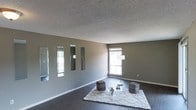 This single-floor space presents a clean, contemporary aesthetic defined by its neutral gray walls, dark hardwood flooring, and bright white trim. The main living area is open and airy, benefiting from natural light streaming in through multiple windows that illuminate the room throughout the day. A textured popcorn ceiling adds a classic touch overhead, contrasting with the sleek, modern elements below. The living space is tastefully appointed with a plush white area rug grounding the seating area, complemented by woven ottomans that add a touch of texture and warmth. A trio of tall, vertical mirrors adorns one wall, strategically placed to reflect light and amplify the sense of openness in the room. A slender arc floor lamp adds a sophisticated lighting element, enhancing the room's modern ambiance. The overall design sensibility leans toward minimalism, allowing the architectural features — clean lines, generous floor space, and well-proportioned windows — to take center stage. The space offers a calm, neutral palette that provides a versatile backdrop for a variety of interior styles and personal touches.