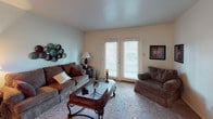 This single-floor residence offers a well-organized layout across several distinct living spaces. The living room serves as the heart of the home, finished with plush carpeting and neutral walls that create a warm, inviting atmosphere. A set of French doors floods the room with natural light and provides access to the outdoors. Adjacent to the living room, a dedicated dining area features a ceiling fan fixture overhead and a charming porthole-style window, adding a unique architectural detail to the space.

The kitchen is designed for practicality, featuring warm wood cabinetry extending to the upper and lower walls, a double-basin stainless steel sink, white appliances including a dishwasher and refrigerator, and light tile flooring. Ample counter space and cabinetry provide generous storage and workspace.

The two bedrooms are carpeted and well-proportioned, each accommodating full-sized furniture comfortably. Both rooms benefit from large windows dressed with horizontal blinds, allowing for controlled natural light. One bedroom includes a built-in open closet with shelving, while the other offers additional storage. The bathroom features a wood vanity with a white countertop, a louvered medicine cabinet, a lighted mirror, and a separate water closet with a shower-tub combination enclosed by a curtain.