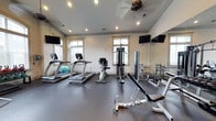 Located at 1001 Bear Island Rd in Summerville, South Carolina, this well-equipped fitness center offers a comprehensive workout environment across a single level. The space is divided into two distinct exercise areas, each thoughtfully designed to support a variety of fitness routines. The first area features a durable rubber flooring surface and is outfitted with treadmills, a weight bench, a cable machine, and free weights, complemented by wall-mounted mirrors that enhance the sense of space. Recessed lighting and crown molding lend a polished, finished look, while multiple windows allow natural light to fill the room. Wall-mounted flat-screen TVs are positioned for easy viewing during workouts. The second area boasts a vaulted ceiling with an arched window as a focal point, creating an airy, open atmosphere. This section includes elliptical trainers, treadmills, a selection of kettlebells, and stability balls, with additional wall-mounted TVs for entertainment. A ceiling fan provides comfortable airflow throughout. Together, both areas provide a well-rounded facility suitable for cardio training, strength building, and functional fitness.