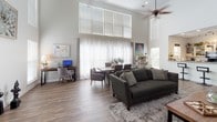 Located at 15235 Oneal Rd in Gulfport, Mississippi, this single-story home impresses with its dramatic vaulted ceilings and thoughtfully designed open-concept floor plan. The expansive main living area serves as the heart of the home, anchored by a stunning floor-to-ceiling natural stone fireplace that commands attention and adds warmth and texture to the space. Wide-plank hardwood-style flooring flows continuously throughout, unifying each area with a cohesive, modern aesthetic.

Natural light pours in generously through oversized windows fitted with sheer curtains and horizontal blinds, creating a bright and airy atmosphere. Ceiling fans are positioned throughout to enhance comfort. The living area transitions effortlessly into a dedicated dining space and a functional workspace nook, making the layout as practical as it is visually appealing.

The kitchen is accessed through a wide arched opening and features a raised breakfast bar with seating, open shelving, track lighting, and black appliances. Its semi-open design maintains visual connection to the main living areas while providing a defined cooking space.

Additional rooms visible from the main living area suggest further living or formal spaces, accessed through elegant French doors. The overall ambiance of the home is welcoming and contemporary, blending casual comfort with distinctive architectural character.