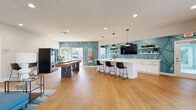 Located at 6158 Beacon Isles Dr in Town N' Country, Florida, this single-floor community space offers a thoughtfully designed and vibrant gathering environment. The interior is unified by warm hardwood-style flooring, recessed lighting, and a cohesive coastal color palette anchored by striking teal geometric accent walls.

The main social hub combines a fully equipped kitchenette and bar area featuring an expansive white quartz island with bar seating, crisp white cabinetry with matte black hardware, open shelving, and pendant lighting. A wall-mounted TV and decorative greenery complete the space, making it ideal for casual gatherings and events.

Adjacent to the kitchen is a spacious game and recreation area, highlighted by a shuffleboard table and a beverage vending machine, with large windows that invite natural light and views of the outdoors.

The lounge area provides a comfortable retreat with a large blue sectional sofa, coordinating accent chairs, a wall-mounted TV, and an area rug with coastal-inspired tones. Large windows and glass doors connect the interior to the outdoor surroundings, reinforcing an open, airy atmosphere.

A separate dining nook features a modern table with a geometric pendant light fixture and expansive sliding glass doors that flood the space with natural light and offer direct views of the outdoor deck and surrounding landscape. Together, these areas create a welcoming, multi-functional community facility.