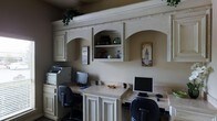This home office is thoughtfully designed to accommodate two users simultaneously, featuring a continuous built-in desk surface that spans the width of the room. Each workstation is equipped with a computer monitor and ergonomic office chair, providing dedicated workspace on either end. A printer station is integrated into the left side of the setup, keeping office essentials organized and accessible.

The cabinetry is a standout architectural element, finished in a creamy antique white with detailed raised-panel doors and decorative rope-style crown molding that runs across the top. Three arched open shelving niches serve as the focal point of the upper cabinetry, offering display space for decorative objects, books, and personal accents. Additional enclosed upper and lower cabinets provide generous storage throughout.

The textured walls in a warm neutral tone complement the cabinetry finish, contributing to a cohesive and inviting aesthetic. Trailing greenery atop the upper cabinets adds a natural touch. Overall, the space balances functional office design with classic, refined detailing.