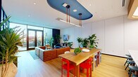 This thoughtfully designed single-floor space makes a striking first impression through its bold, eclectic aesthetic and open-plan layout. The centerpiece of the room is a distinctive oval blue ceiling panel suspended above the dining area, from which sleek linear pendant lights hang on leather cord suspensions, creating an artistic focal point that anchors the space. Light oak hardwood flooring runs throughout, lending warmth and continuity to the open floor plan.

The dining area features a solid wood communal table surrounded by a lively mix of multicolored chairs in red, purple, blue, and green — an intentional design choice that adds energy and personality. Adjacent to the dining area is a curved kitchen counter with rounded edges, backed by a vivid, patterned mosaic tile wall in warm reds and oranges, providing a bold artistic backdrop.

The lounge area is anchored by a substantial ribbed leather sectional sofa, complemented by an array of eclectic seating including a blue accent chair and a patterned lounge chair, all arranged around a round lacquered coffee table atop a blue geometric area rug.

Floor-to-ceiling glazed panels and French doors along one wall flood the space with abundant natural light while offering direct access to an outdoor area. Lush indoor tropical plants are placed throughout, adding a vibrant, biophilic dimension to the overall design. The space is further defined by clean white panel walls that balance the room's colorful, layered character.