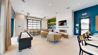 Located at Yardly Artisan Lakes in Palmetto, Florida, this single-floor resident clubhouse delivers a sophisticated yet relaxed gathering space designed for community living. The open-concept layout is anchored by a central lounge area furnished with woven rattan armchairs, ottomans, and a round marble-top coffee table atop a layered area rug, all positioned in front of a sleek linear electric fireplace topped by a wall-mounted TV. A striking deep navy accent wall provides bold contrast against the bright white textured walls and light-toned large-format tile flooring throughout.

A dedicated bar and kitchenette area features warm wood cabinetry, quartz countertops, a beverage refrigerator, and open shelving adorned with a live moss wall backdrop and neon signage. An industrial-style glass garage door opens to bring in abundant natural light and connects the interior to the bar counter seating. A shuffleboard table adds recreational appeal, while a banquette dining nook with local coastal artwork creates a casual café-like corner.

Large sliding glass doors along one wall flood the space with natural light and open directly onto an outdoor terrace with views of the resort-style pool, palm trees, and lounge seating — seamlessly blending indoor comfort with Florida's outdoor lifestyle.