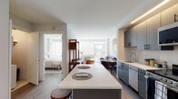 This single-floor residence at 50 Lecount Place in New Rochelle, NY, offers a thoughtfully designed open-concept layout with modern finishes throughout. The kitchen serves as a central highlight, featuring matte gray cabinetry with black hardware, a white quartz countertop, a stainless steel undermount sink, and a full suite of stainless steel appliances including a range, microwave, dishwasher, and refrigerator. A substantial island with a white quartz surface and bar seating provides additional workspace and casual dining. Recessed linear lighting above the cabinetry enhances the clean, contemporary aesthetic, while light wood-tone flooring runs throughout, unifying the space. The open living and dining area flows naturally from the kitchen, with large windows drawing in natural light and providing outdoor views. A compact dining table and seating area complement a comfortable sofa arrangement. The bedroom is calm and understated, featuring neutral tones, a closet, and soft natural light. The bathroom is finished with large-format tile flooring, a dark vanity with a white countertop, a framed mirror, and contemporary fixtures, creating a clean and polished environment overall.