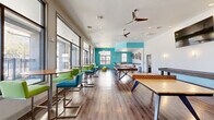 Located at 2002 Stambuk Ln in Statesboro, Georgia, this single-story community clubhouse spans a generously sized footprint and is designed to serve a variety of social, recreational, and functional needs. The interior is characterized by warm hardwood-style flooring, crisp white walls accented with bold teal highlights, and recessed lighting complemented by ceiling fans throughout.

The main gathering area features floor-to-ceiling windows that flood the space with natural light and offer views of an outdoor pool and surrounding grounds. This open zone houses a pool table with hairpin legs, a shuffleboard table, and high-top seating with vibrant green and teal chairs, creating an energetic game and lounge environment. An adjacent seating area includes a gas fireplace, large mounted television, decorative pendant lighting, and a teal coffered ceiling detail, providing a comfortable social lounge.

A dedicated computer lab offers built-in workstations along teal-painted walls with ample natural light. A private study room features a round table, yellow task chairs, and a full whiteboard. The kitchenette includes white cabinetry, dark countertops, and decorative metallic wall art. Additional amenities include a tanning room with a tanning bed, full-length mirror, and clothes storage.
