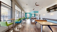 Located at 2002 Stambuk Ln in Statesboro, Georgia, this single-story community clubhouse spans a generously sized footprint and is designed to serve a variety of social, recreational, and functional needs. The interior is characterized by warm hardwood-style flooring, crisp white walls accented with bold teal highlights, and recessed lighting complemented by ceiling fans throughout.

The main gathering area features floor-to-ceiling windows that flood the space with natural light and offer views of an outdoor pool and surrounding grounds. This open zone houses a pool table with hairpin legs, a shuffleboard table, and high-top seating with vibrant green and teal chairs, creating an energetic game and lounge environment. An adjacent seating area includes a gas fireplace, large mounted television, decorative pendant lighting, and a teal coffered ceiling detail, providing a comfortable social lounge.

A dedicated computer lab offers built-in workstations along teal-painted walls with ample natural light. A private study room features a round table, yellow task chairs, and a full whiteboard. The kitchenette includes white cabinetry, dark countertops, and decorative metallic wall art. Additional amenities include a tanning room with a tanning bed, full-length mirror, and clothes storage.