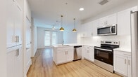 This thoughtfully designed one-bedroom, one-bathroom residence in Austin, TX offers a clean, modern aesthetic across its single-floor layout. The open-concept living and kitchen area is a standout feature, anchored by a peninsula island with an undermount sink and crisp white quartz countertops. White shaker cabinetry lines the kitchen walls, complemented by stainless steel appliances including a range, built-in microwave, dishwasher, and refrigerator. Two gold dome pendant lights hang above the island, adding a warm, contemporary accent against the bright white interior. Light wood-tone flooring runs throughout the main living area, enhancing the airy, open feel of the space. Large windows and a glass door at the far end of the living area invite abundant natural light and provide outdoor access. The bedroom is a comfortable, well-proportioned retreat finished with soft carpeting and a generously sized window dressed with blinds, while a ceiling fan adds practical comfort. The bathroom continues the clean, modern palette with a wide vanity featuring white cabinetry, a quartz countertop, a large mirror, and wood-look flooring. A dedicated closet completes the unit, offering organized storage. The overall ambiance is bright, fresh, and move-in ready.