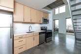 Located at 219 E Grand Ave in the heart of Des Moines, Iowa, this two-story loft offers a modern, industrial-inspired design across two well-proportioned floors. Polished concrete floors run throughout, lending a clean, contemporary aesthetic that ties the space together cohesively.

The main floor hosts the kitchen, which is outfitted with light maple cabinetry, white countertops, stainless steel appliances including a refrigerator, range, over-the-range microwave, and dishwasher, along with a single-basin sink. The open layout of the main floor is bathed in natural light from large windows and a skylight, with direct views of the surrounding Des Moines cityscape. A striking spiral staircase with an industrial metal frame connects the two levels.

The upper floor features an open loft area with exposed ductwork along the ceiling, recessed lighting, and a perforated metal railing that overlooks the main floor below. Large windows on this level provide continued urban views and abundant daylight.

The two bathrooms are finished in a clean, modern style, with one featuring mosaic tile surrounds, a soaking tub, and a wall-mounted sink. Additional conveniences include a dedicated closet with wire shelving and a hallway connecting the functional areas of the space. The overall ambiance is minimalist and urban, well-suited for a live-work lifestyle in downtown Des Moines.