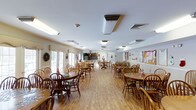 This single-floor space is designed to serve as a communal gathering and dining area, offering a warm and inviting environment for its occupants. The main hall is generously proportioned, accommodating numerous round wooden tables paired with classic Windsor-style chairs arranged throughout the open floor plan. Light hardwood flooring runs the length of the room, contributing to the space's bright and cheerful character.

Natural light filters in through several windows and glass-paneled doors along one wall, complementing the flush-mounted ceiling light fixtures that keep the interior well illuminated. A service counter or kitchenette area is visible toward the back of the room, suggesting convenient on-site food and beverage service capabilities.

The walls are adorned with bulletin boards, decorative displays, and seasonal holiday décor, adding a lively, community-centered ambiance. HVAC units are mounted on the ceiling to ensure year-round comfort. The layout is thoughtfully organized to allow easy movement between tables, making it well-suited for group dining, social events, or organized activities. Overall, the space strikes a balance between functional design and a welcoming, homey atmosphere ideal for community gatherings.
