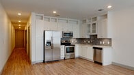 This single-floor unit offers a thoughtfully designed layout with a seamless flow between the living and kitchen areas. The open-concept living space is finished with warm-toned hardwood flooring and recessed lighting, creating a bright and inviting atmosphere. Large windows allow natural light to fill the room, enhancing the clean, neutral color palette of white walls and light trim.

The kitchen is a standout feature, boasting crisp white shaker-style cabinetry that extends to the ceiling with open display shelving above. A full suite of stainless steel appliances — including a side-by-side refrigerator with ice maker, electric range, over-the-range microwave, and dishwasher — is complemented by a grey subway tile backsplash and light-toned countertops. The layout maximizes both storage and workspace efficiently.

A hallway connects the living areas to the private spaces, featuring louvered double doors that provide access to a laundry or storage closet, alongside a sleek barn-style sliding door. The bathroom is well-appointed with grey cabinetry, a wide vanity, white subway tile surrounding the shower-tub combination, and large-format tile flooring. The overall design maintains a cohesive, contemporary aesthetic with neutral tones and quality finishes throughout.