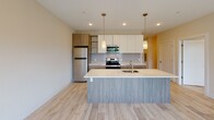 This thoughtfully designed one-bedroom, one-and-a-half-bathroom residence with a den is located at Somerville Parc in Somerville, NJ. The single-floor layout offers a comfortable and functional living experience with modern finishes throughout.

The open-concept kitchen serves as a central highlight, featuring a large island with an undermount sink, light quartz countertops, and a blend of white upper cabinetry and wood-toned lower cabinets. Stainless steel appliances, a tile backsplash, and stylish pendant lighting complete the contemporary design. Light-toned hardwood-style flooring flows seamlessly through the main living areas, enhancing the bright, airy ambiance.

The spacious living area benefits from large windows and a glass door providing direct access to a private balcony with outdoor views of lush greenery. An additional room serves as a versatile den or bedroom, also featuring a large window with exterior views. The primary bathroom includes a glass-enclosed walk-in shower with textured tile surround and wood-look tile flooring, while the half-bath offers a wall-mounted vanity and large mirror. Recessed lighting and neutral warm-toned walls tie the entire space together with a clean, modern aesthetic.