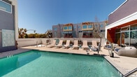 Located at 160 S Ventura Ave in Ventura, California, this outdoor amenity deck offers a well-appointed communal retreat designed for relaxation and leisure. The centerpiece of the space is a generously sized swimming pool with crystal-clear turquoise water, surrounded by a broad sun deck finished in light-toned concrete that reflects the abundant Southern California sunshine. A row of sling-style lounge chairs lines the poolside, providing ample space for sunbathing beneath the consistently clear blue skies of the Ventura coast. Flanking the pool area is a dedicated lounge zone featuring distinctive wicker pod daybeds — curved, canopy-style seating units cushioned in striped fabric and accented with colorful throw pillows and small side tables. These semi-enclosed pods offer a comfortable shaded retreat from the sun. The surrounding building architecture is contemporary and multi-story, featuring a mix of gray panel siding, warm wood accents, and large windows that overlook the pool deck. The broader complex context includes glimpses of palm trees and neighboring rooftops, underscoring the property's coastal California setting. The layout is open and airy, with distinct zones for active swimming and quiet lounging, making it a versatile outdoor amenity space suited for a residential community.